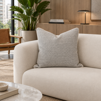 Cojín gris jaspeado con borde claro sobre sofá crema en un living luminoso con madera y mármol