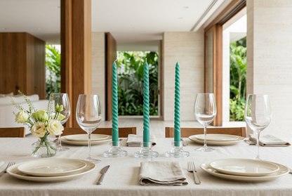 Velas Twister verdes en portavelas de vidrio sobre una mesa elegante con platos marfil, copas y flores discretas.