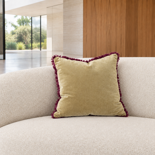 Cojín velvet verde con flecos burdeo sobre sofá marfil en un ambiente moderno y luminoso